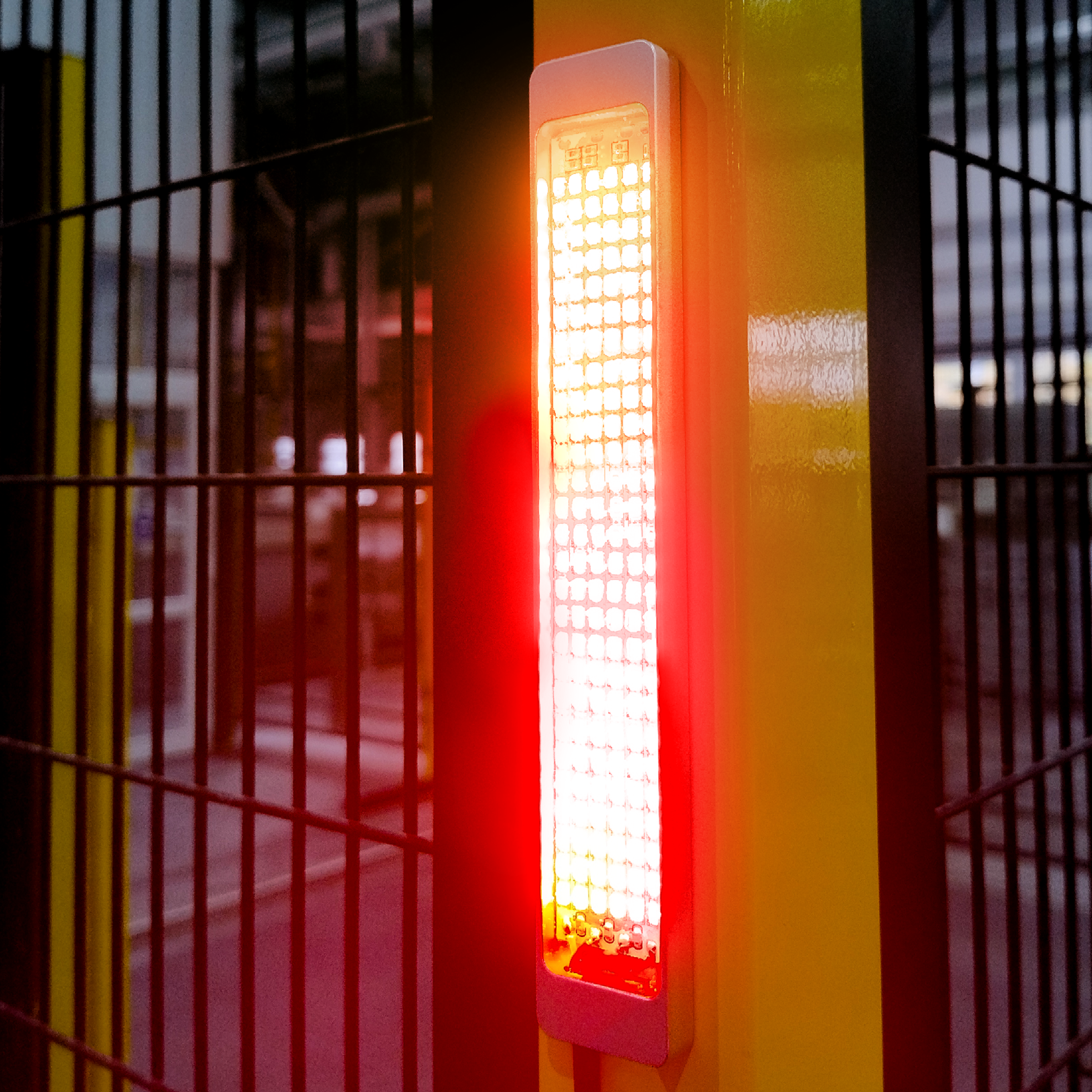 Rectangular red LED signal light glowing on a yellow vertical post next to a black metal fence with vertical bars.