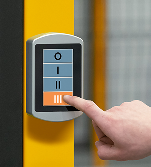 Hand pressing the bottom button labeled 'III' on a rectangular control panel with four buttons mounted on a yellow surface