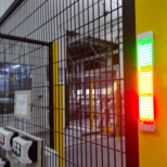 Vertical industrial indicator light with green, yellow, and red lights mounted on yellow post next to a wire mesh fence inside an industrial facility