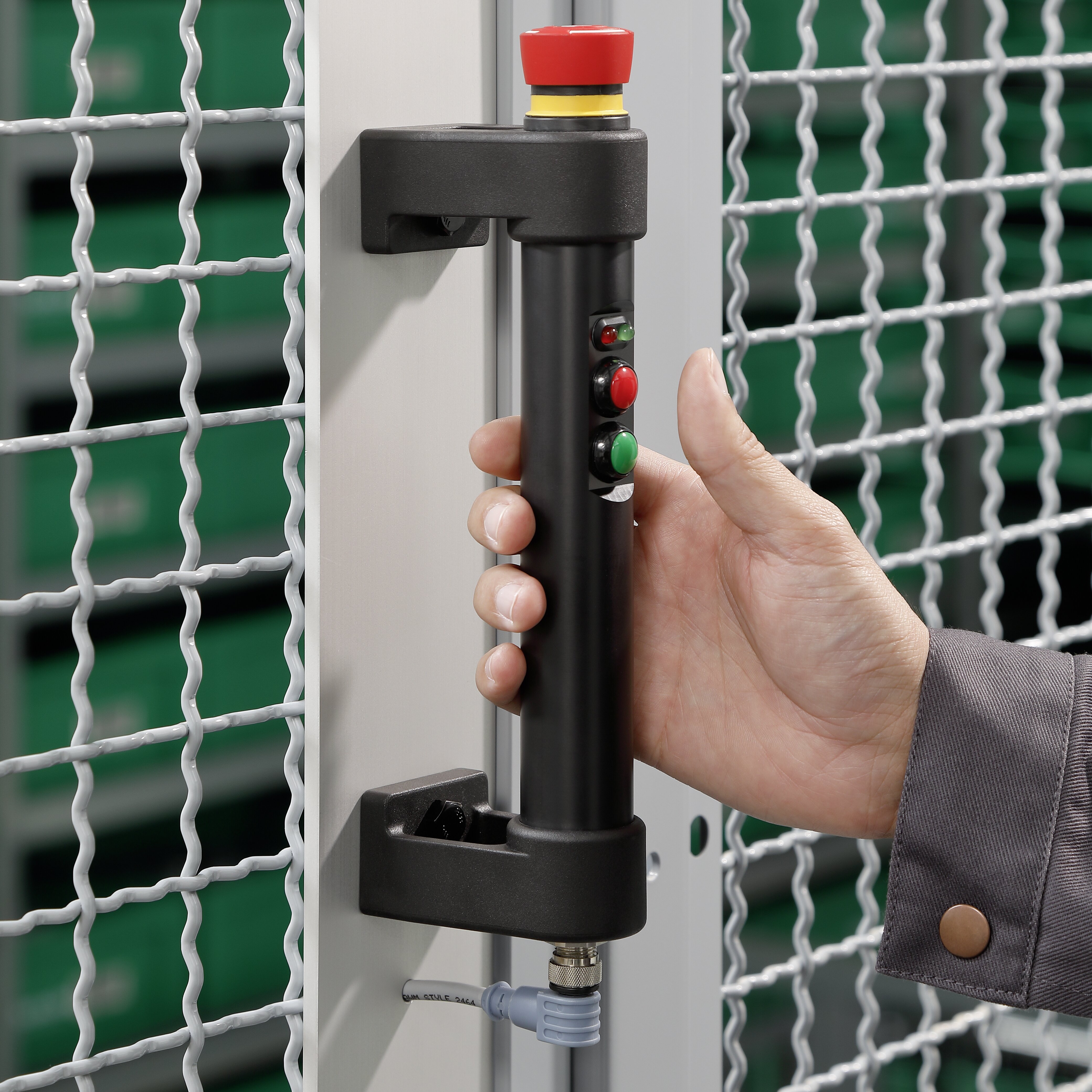 Hand greift schwarzen Griff mit roten und grünen Tasten, befestigt an metallischem Zaun mit grauem Kabelanschluss