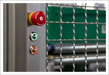 Close-up of an industrial safety panel with a red emergency stop button, green and orange illuminated control buttons next to a metal wire protective fence