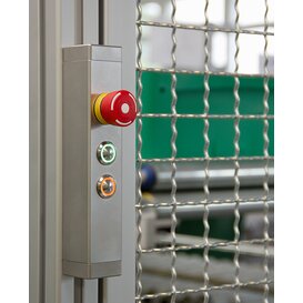 Stainless steel switch housing mounted on a frame with a large red emergency stop button and two illuminated push buttons, next to a metal safety fence with a grid pattern.